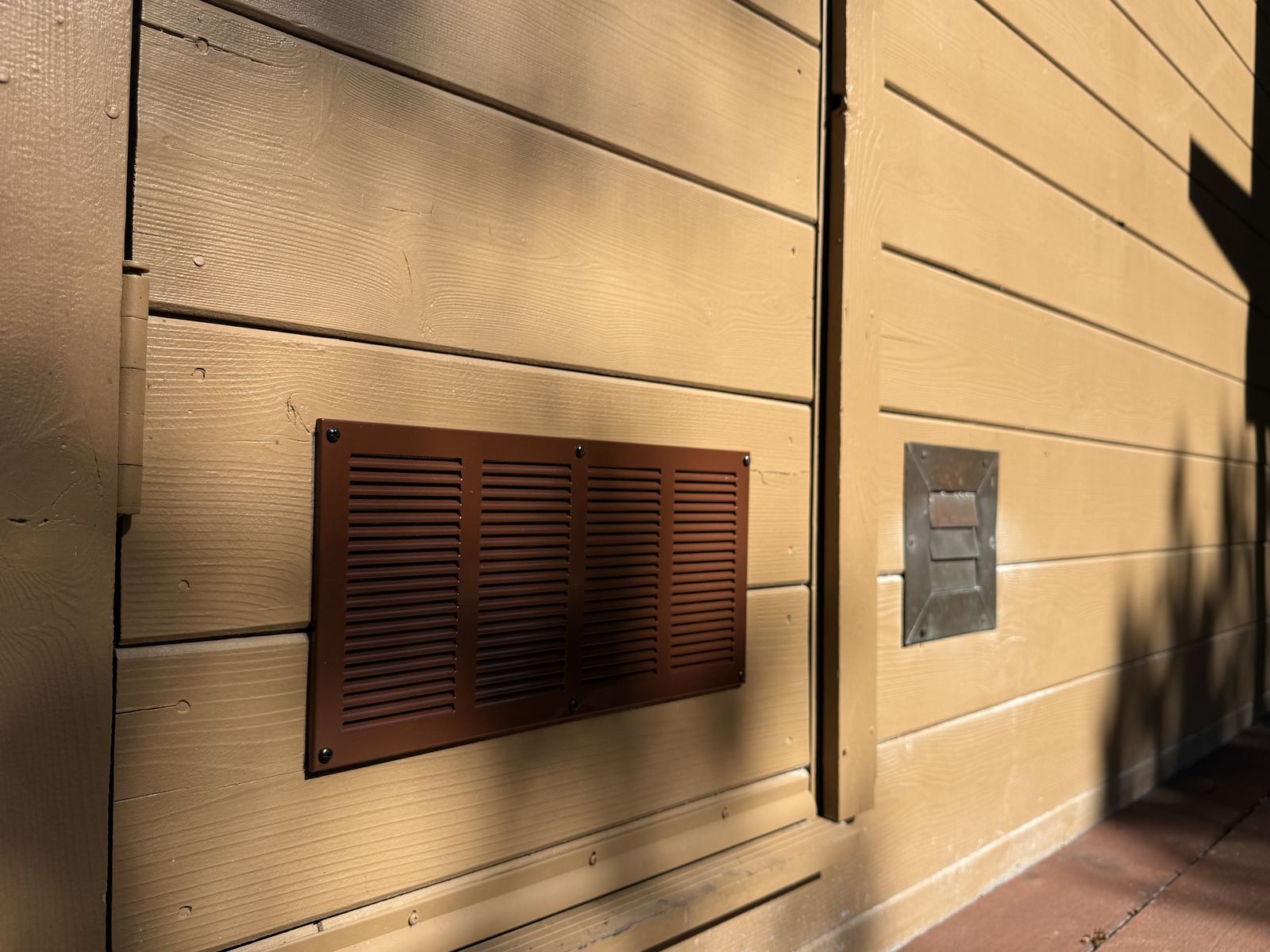 New brown louvered metal vent installed beside an older hooded vent on wood siding